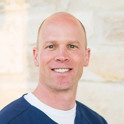 Chris Price smiling in front of a stone wall. He is wearing a blue shirt.
